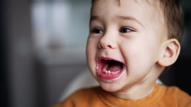 Adorable little kid opens wide his mouth showing few teeth. Baby boy takes a bagel to bite and then is given a spoon of food. Close up.