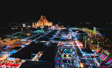Aerial view of beautiful decorative lighting on Thai temple and lots of booths, amusement park, thrilling plaything with crowd of people in annual festival area at night in Samut Sakhon, Thailand