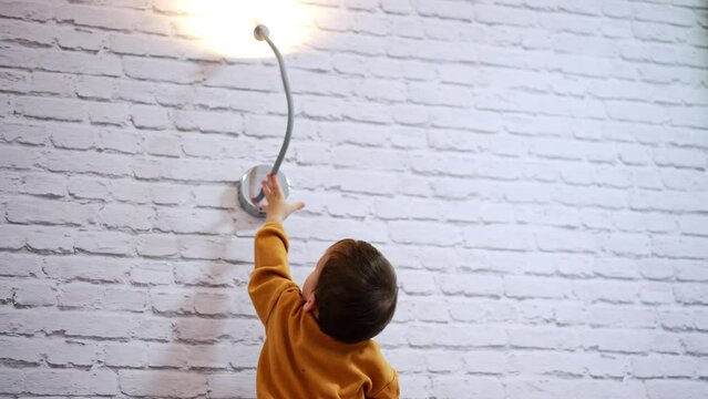 Stubborn baby boy tries to reach the lamp on the wall. Kid cannot grab the illuminator so goes down on the floor. Low angle view.