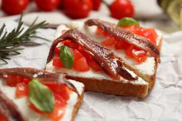 Delicious sandwiches with cream cheese, anchovies, tomatoes and basil on parchment paper, closeup