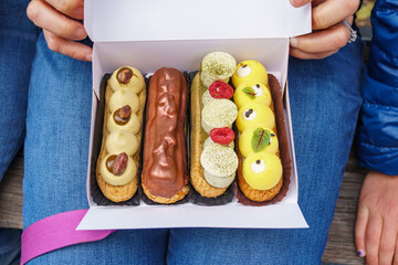 A woman holds a box on her lap with a variety of gourmet eclairs inside