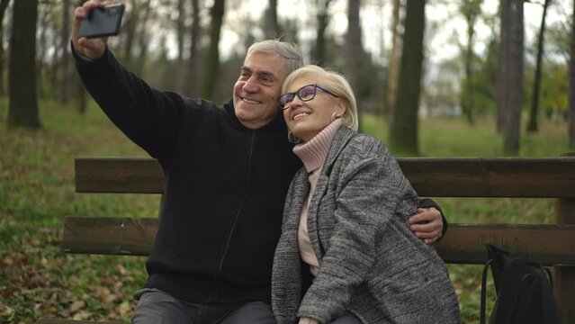 Adult couple sits on a bench in a forest park and takes a selfie together on a smartphone in slow-motion