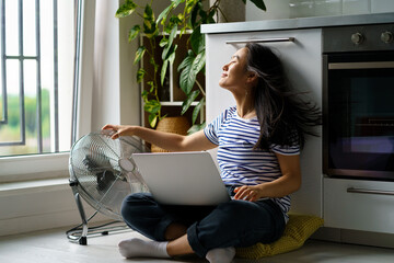 Satisfied Asian woman sits on floor with laptop on crossed knees, enjoying wind from electric fan....