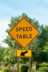 Road sign that says speed table with black and yellow paint on exterior in late afternoon shade in summer sunlight