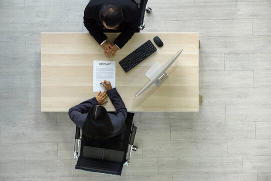 Young candidate in gray suit put a sign on the contract document after being considered by Human Resource Manager. Computer monitor, keyboard and mouse are on wooden table. Top View