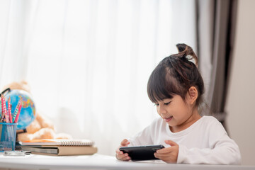 Asian little girl  using a phone close up, distracted from studying, sitting at a table with notebooks, a pretty child having fun with a smartphone, watching the webinar, homeschooling