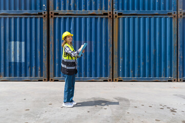 Young caucasian woman with safety vest and yellow hardhat looking at container storage location while holding clipboard. A large cargo container is in the background.