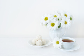 Fototapeta na wymiar Cup of tea, sweets, bouquet or chamomile or daisy flowers on a white wooden table. Tea and flowers on a white background