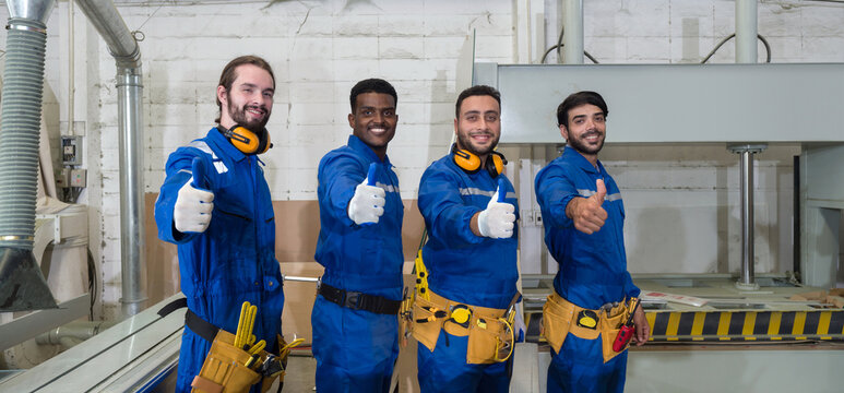 Group of technician in blue mechanic jumpsuit, ear muff and yellow tool belts stand with their thumbs up in front of polywood cold press machine. Production line atmosphere in furniture factory