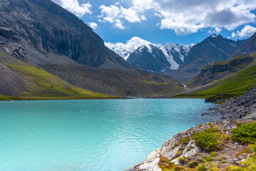 Mountain lake with turquoise water Karakabak in the Altai mountains with snowy peaks and glaciers with under the clouds and green grass with a hanging stone.