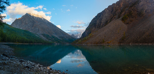 Panorama of the shore of Lake Shavlinskoe with smoke from a tourist fire in the shade among the mountains with reflection of peaks with glaciers and snow in Altai.
