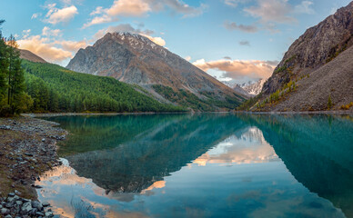 Fototapeta na wymiar Panorama of the turquoise transparent lake Shavlinskoe in the shade with stones among the mountains with reflection of the peaks with glaciers and snow in Altai.