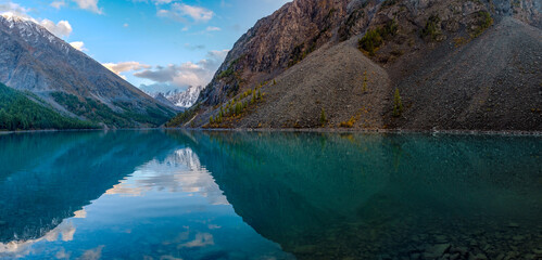 Panorama of the turquoise transparent lake Shavlinskoe in the shade with stones among the mountains with reflection of the peaks with glaciers and snow in Altai in the evening.