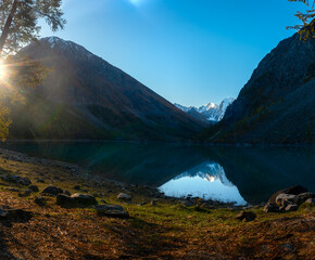 Panorama of Lake Shavlinskoe in the shade with stones among the mountains with reflection of the peaks with glaciers and snow in Altai in the evening with the rays of the sunset.
