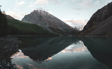 Panorama of lake Shavlinskoe in the shade with stones among mountains with reflection of peaks with glaciers and snow in Altai in the evening.