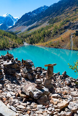 Stones stacked in pillars on the steep shore of the high-altitude turquoise lake Shavlinskoye against the backdrop of mountains with glaciers and snow in Altai.
