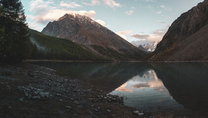 Shore with clear water of Lake Shavlinskoe with stones and dry autumn grass in Altai against the backdrop of mountains with snow and glaciers in the evening at dusk.