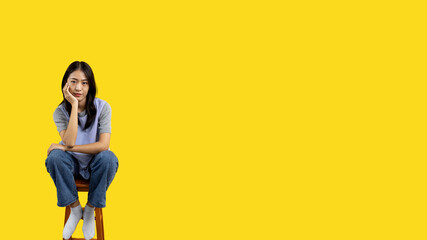 Asian biracial woman sitting in wooden chair and smiling happily on yellow background, Lovely...