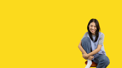 Asian biracial woman sitting in wooden chair and smiling happily on yellow background, Lovely...