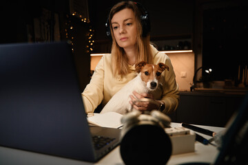 Woman sitting at table with her dog and looking in laptop screen. Female freelancer working from home office at night with pet.