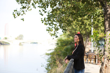 Beautiful young woman leaning over the railing of the river in Seville, Spain. The woman is happy and relaxed looking at the horizon where you can see the other side of the river with the city.