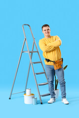 Young man with ladder and cans of paint on blue background