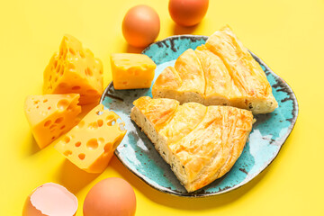 Plate with pieces of delicious cheese pie and ingredients on yellow background