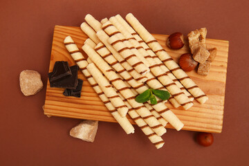 Wooden board with tasty wafer rolls, dark chocolate, nuts and mint on brown background