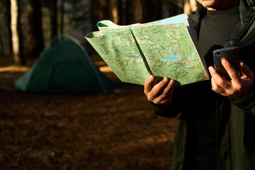 Adult hiker with backpack using mobile phone and map in forest near his tent. Hiking elderly man in autumn nature holding map outdoors next to a campsite in the sunlight closeup shot. Ageless travel