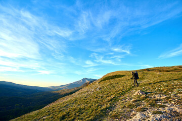 Silhouette of a man in the mountains. Successfully achieving goals. Male hiker on mountain peak with green grass looking at beautiful mountain valley at sunset against clear blue sky.. 