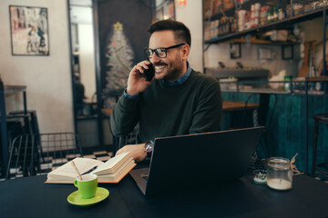 Fototapeta na wymiar Smiling businessman in eyeglasses using laptop and talking on phone in cafe