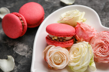 Heart-shaped plate with tasty macaroons and rose flowers on black background. Valentine's Day celebration