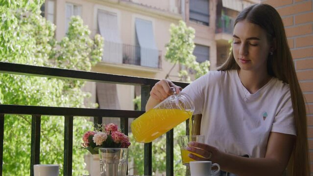 Portrait of young girl in her 20s pouring fresh orange juice into a glass outdoors. Close-up view of blonde-hairs female teenager enjoying tasty breakfast. High quality 4k footage