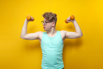 young non-athletic guy with glasses raises small dumbbells on a yellow background and shows surprise