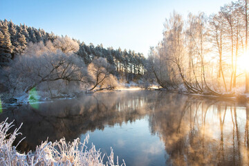 Amazing scenery of river and edge of forest in frosty morning. Ligth mist over river. Trees are reflected on the surface of the river as in the mirorr. Morning sunbeams red color on trees tops. 