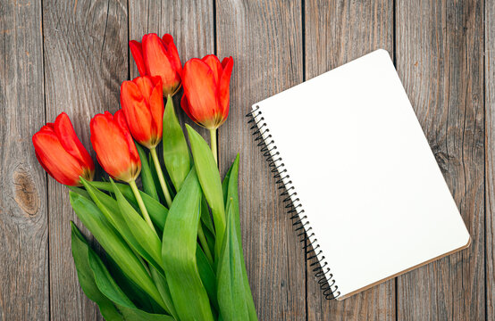 Bouquet of red tulips and notebook on a wooden background, top view.