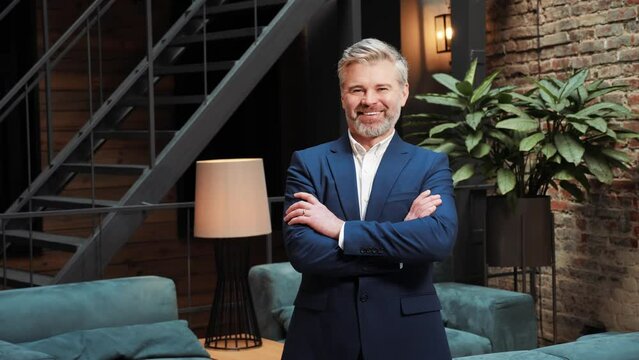 Portrait of confident berded middle age businessman turning head to camera and smiling while crossing arms proud entrepreneur enjoying successful startup company in a modern loft meeting room. 