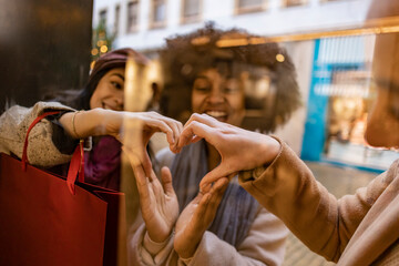Happy young friends making heart shape together with hands seen through glass