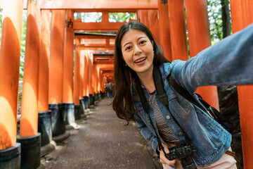 self portrait of happy asian Japanese female tourist smiling at camera while taking pictures with red paralleled senbon torii gates on fushimi inari pathway in Kyoto japan