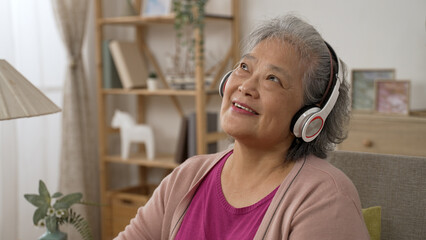 happy chinese grey haired senior lady is smiling and bobbing head with the rhythm while enjoying music from earphones in the living room at home.
