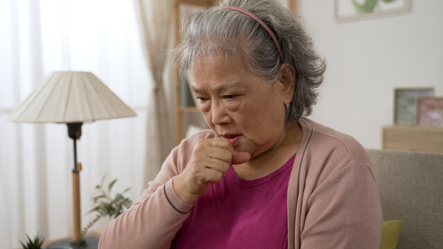 closeup view of an unwell asian grandma getting lung infection is patting on her chest while coughing in the living room at home