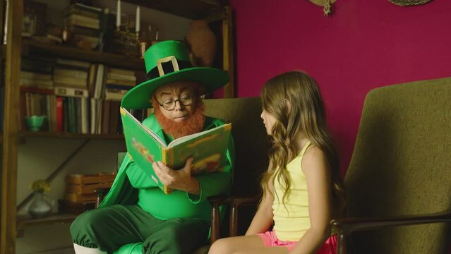Old leprechaun is reading book , telling fairy tale story for little girl . Kid listening carefully and smiling . Many books on background .  Gnome wearing big green hat and traditional costume 