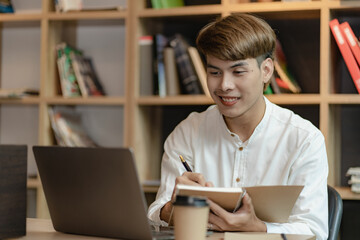 Serious young asian man working in office at desk. Holding and looking at document. Graph. Planning in office. calculation report Using laptop and calculator, online meeting concept