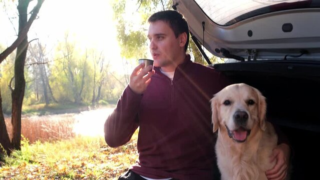 A happy man and his dog are sitting in the trunk of the car drinking hot tea. A young man with a golden retriever on a car trip in the fall woods.