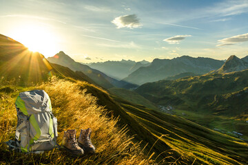 Hiking equipment in early morning sunrise and mountain silhouettes. Backpack and boots in beautiful sunlight and sunbeams. Tyrol, Lechtal, Austria. - 566191305