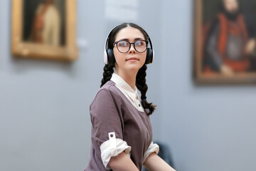 Portrait of young Caucasian pretty woman wearing headphones and contemplates ancient artefacts. Student visiting arts exhibition. Concept of culture education
