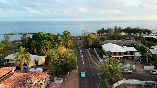 Drone aerial of blue teal retro car  down long winding road along coastline. Past residential neighbourhood, Nightcliff jetty. Native Australian green trees skyline. Sunset Thunderbird car