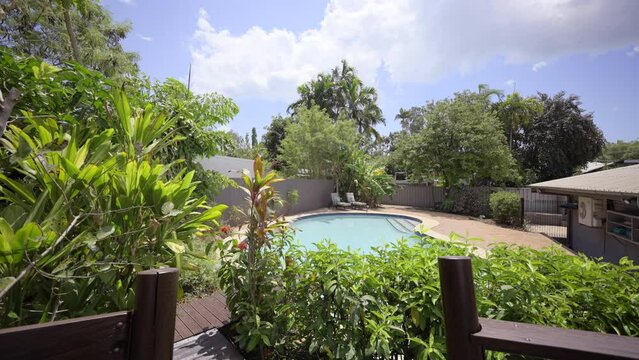 Wooden Deck and rail to outdoor pool. Concrete tiled patio veranda outdoor area. Luxurious pool coping edging. Tropical garden green palm trees. Outdoor sun lounge deck chairs.