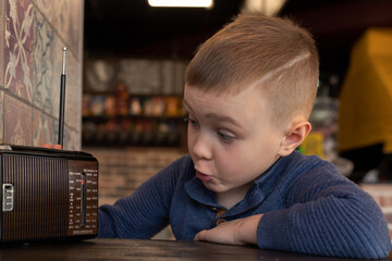 Cute little boy at cafe table with old vintage radio. He is looking at the radio with awe and...