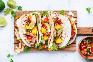 Tacos with grilled chicken, tomato salsa sauce, mango, cilantro and onion in tortilla on cutting board. White table background, top view